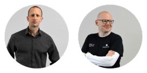 Two men standing side by side, one in a black shirt and the other in a black tee with logos, against a plain background.